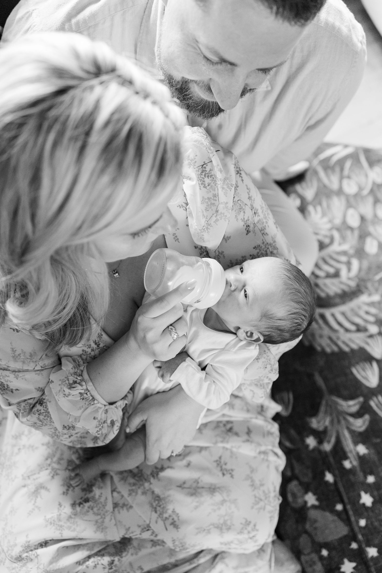 Black and white photo of parents bottle feeding their newborn baby together in a tender moment.