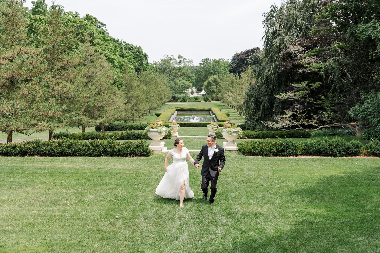A bride and groom hold hands while walking across a lush green lawn in an elegant formal garden setting.