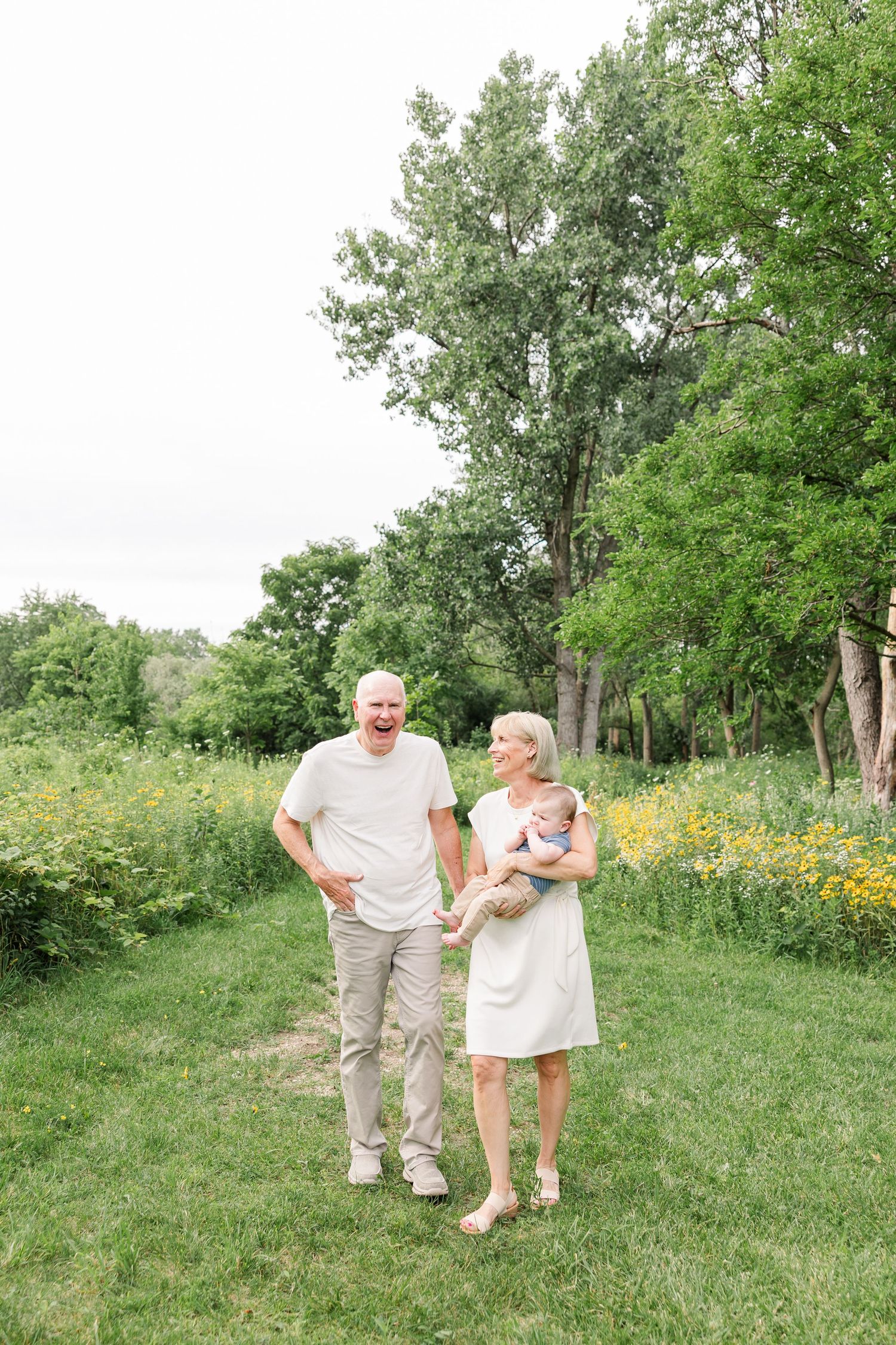 A cheerful older couple walks through a lush green park, grandmother tenderly holding a baby.