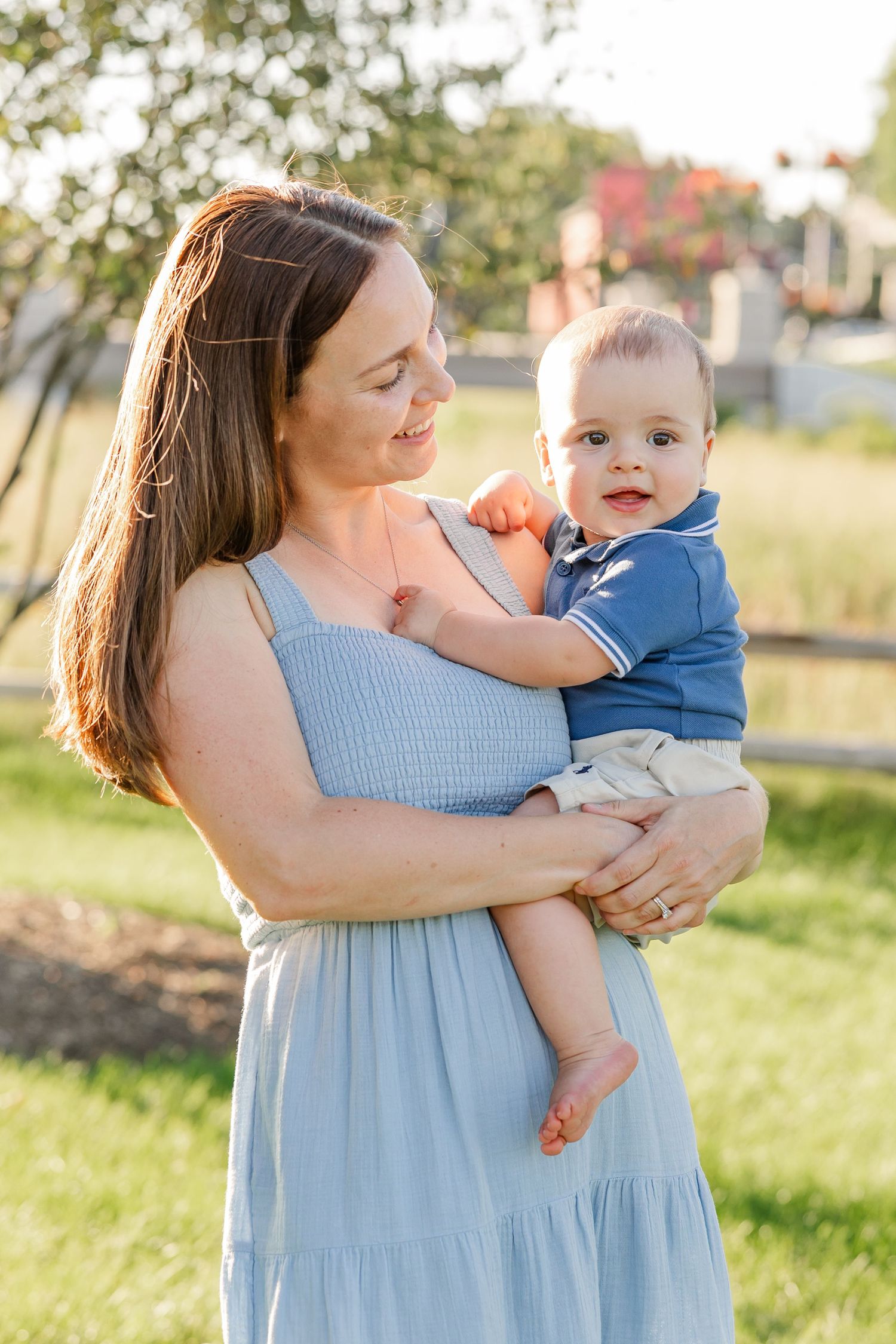 A smiling mother in a blue dress holds her alert baby boy outdoors in warm golden sunlight.