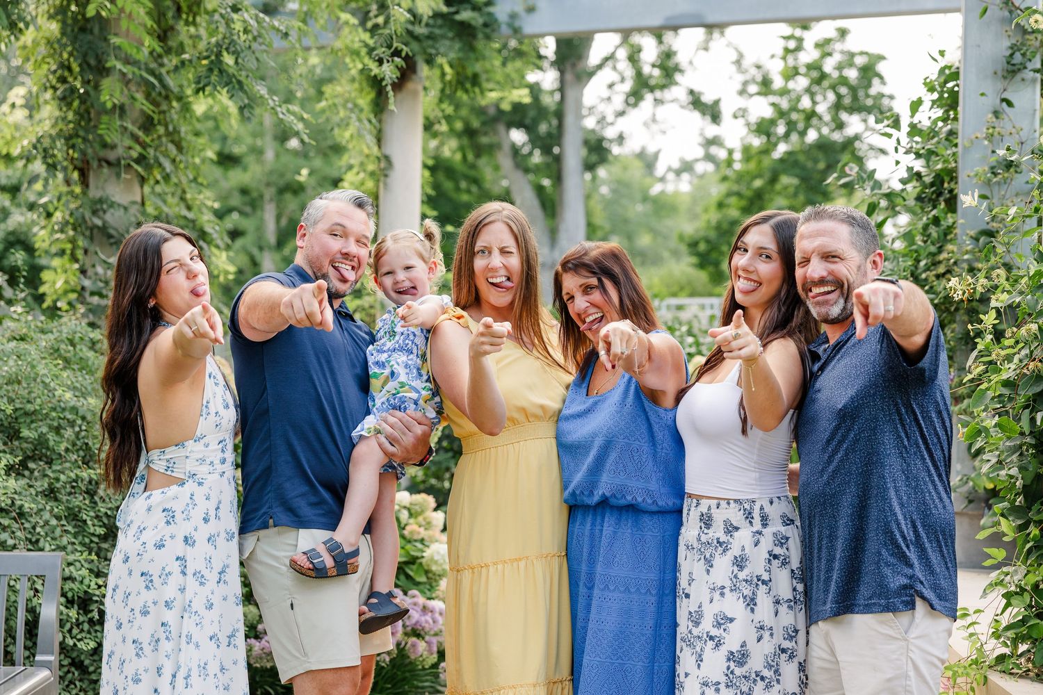Joyful extended family group of seven pointing and laughing together under garden pergola.