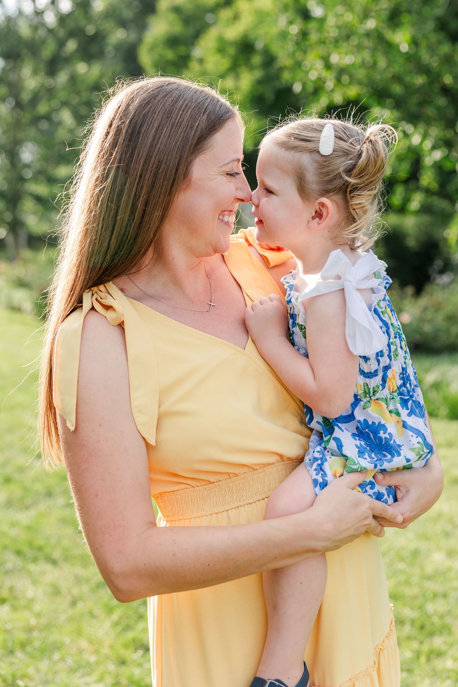 Mother in yellow dress lovingly holding her young daughter nose-to-nose in a sunny park.