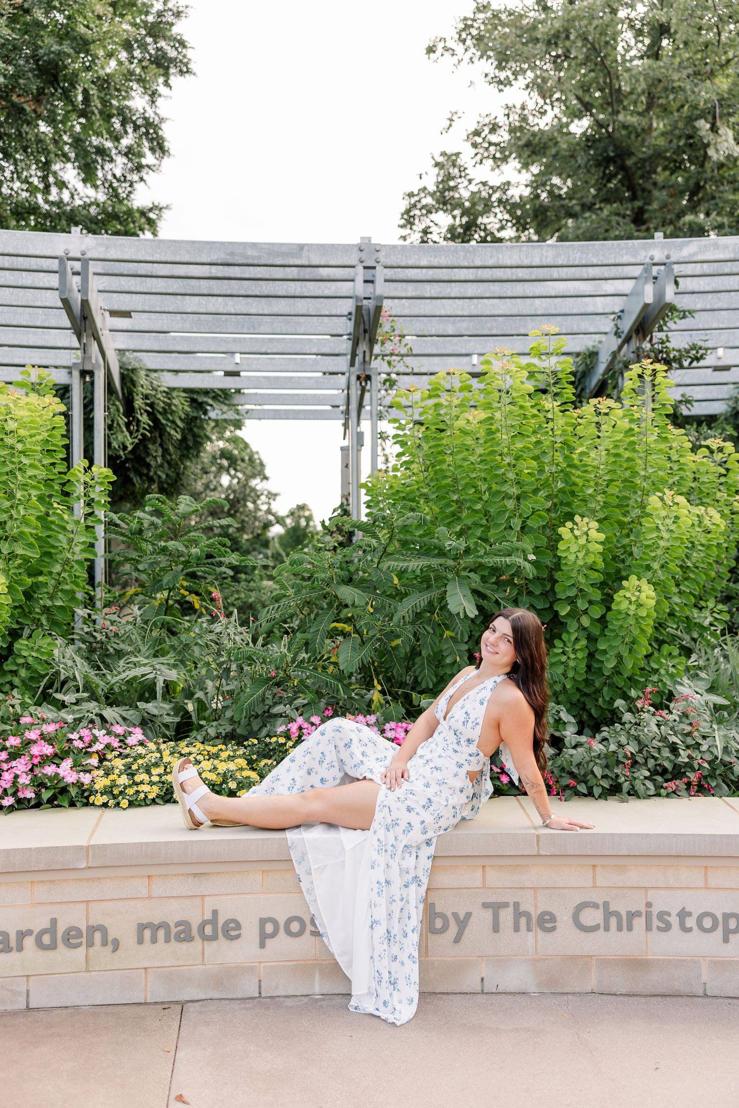 Woman in floral jumpsuit posing on stone wall surrounded by lush garden greenery and colorful flowers near a wooden pergola.