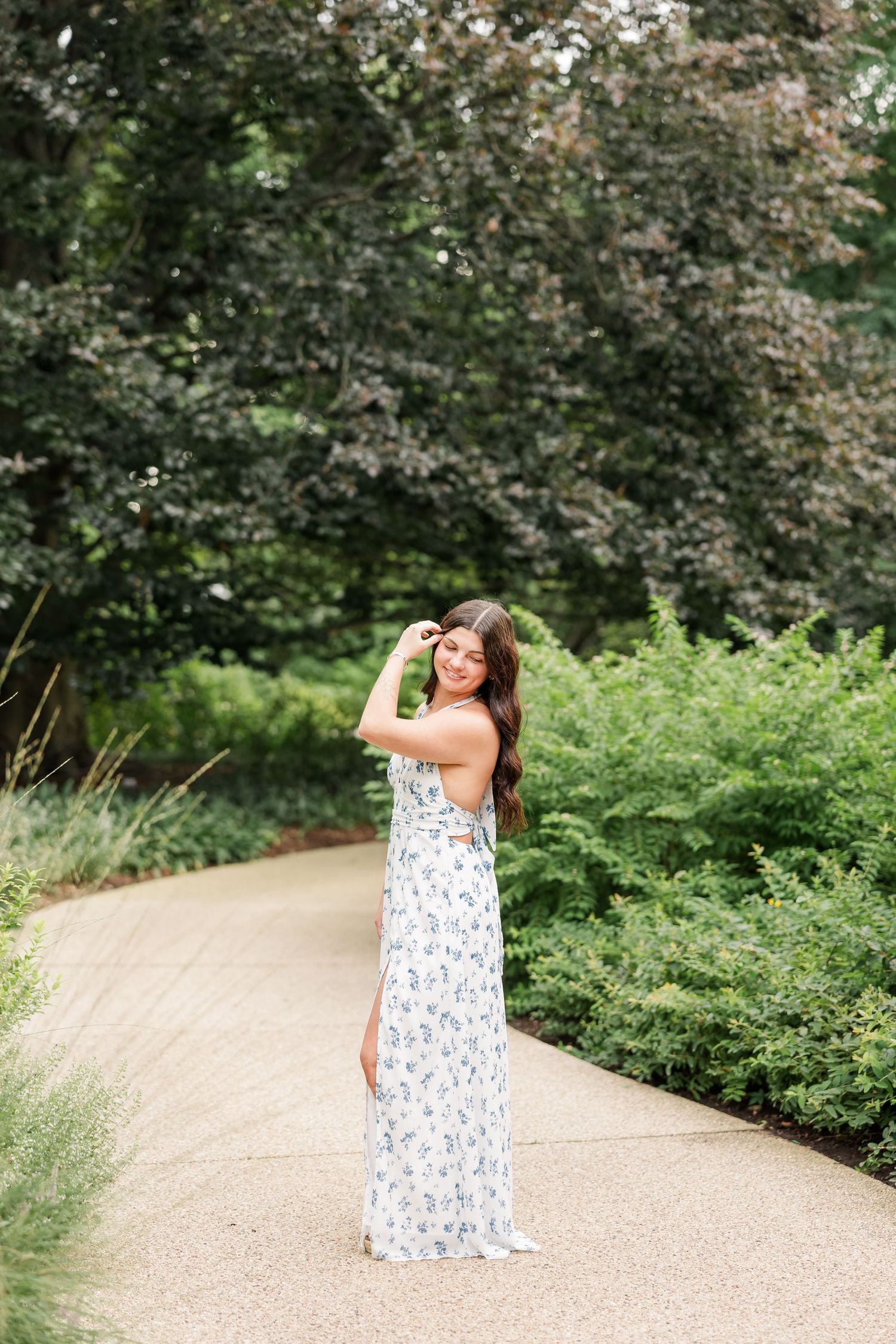 Young woman in white floral maxi dress poses on garden path surrounded by lush green trees and shrubs.