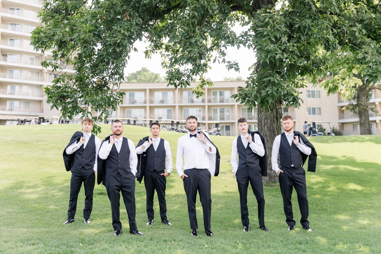 Six groomsmen in black suits and white vests pose casually under a large tree at an outdoor wedding venue.