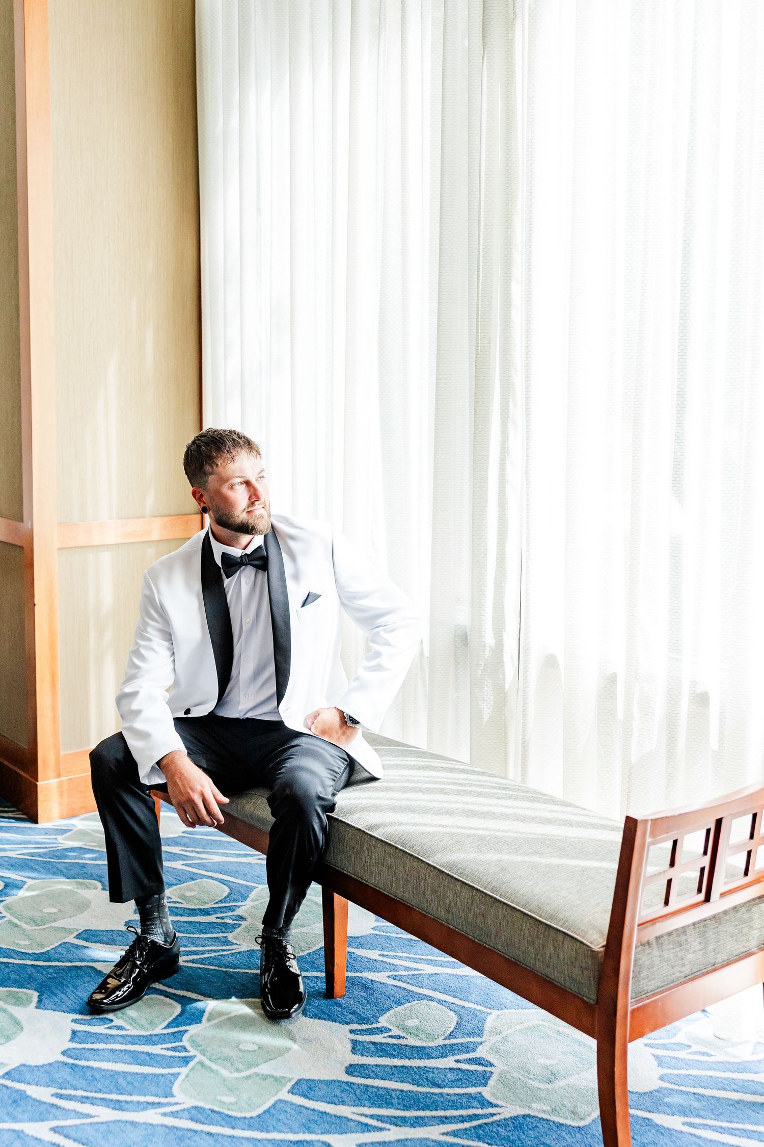 Dapper groom in white tuxedo jacket and black bow tie sitting on bench by hotel window.