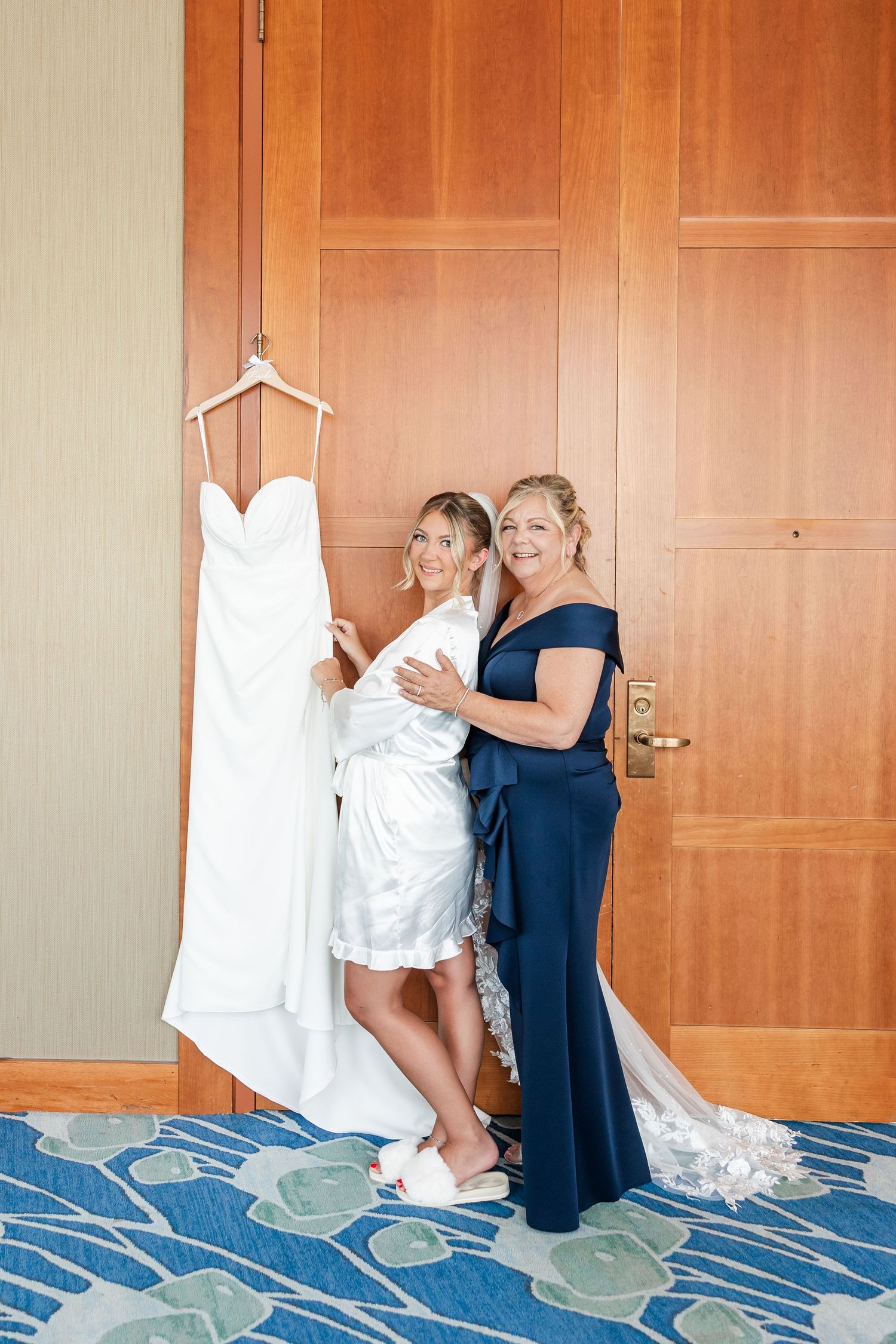 Bride in white robe poses with mother in navy gown beside hanging wedding dress in hotel room.