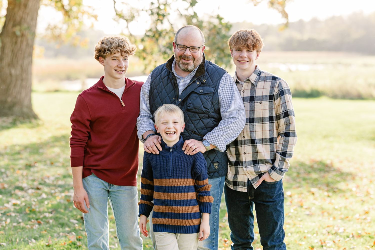 Smiling man in a navy vest with three boys in casual sweaters and plaid shirts posing for a family portrait in a sunny grassy park.