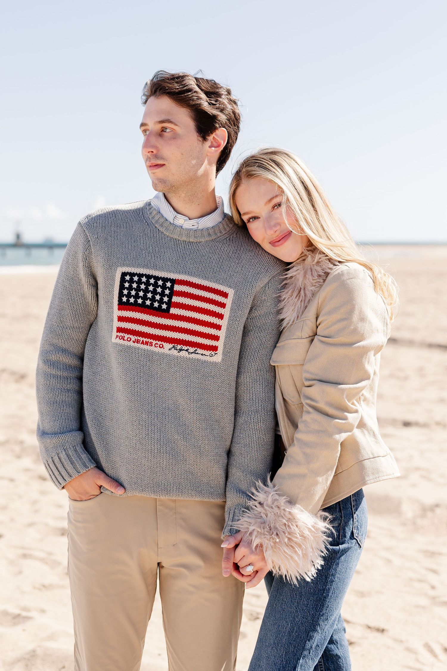 Couple embracing on a sandy beach, man wearing a Polo Jeans Co. American flag sweater, woman in a beige fur-trim jacket.