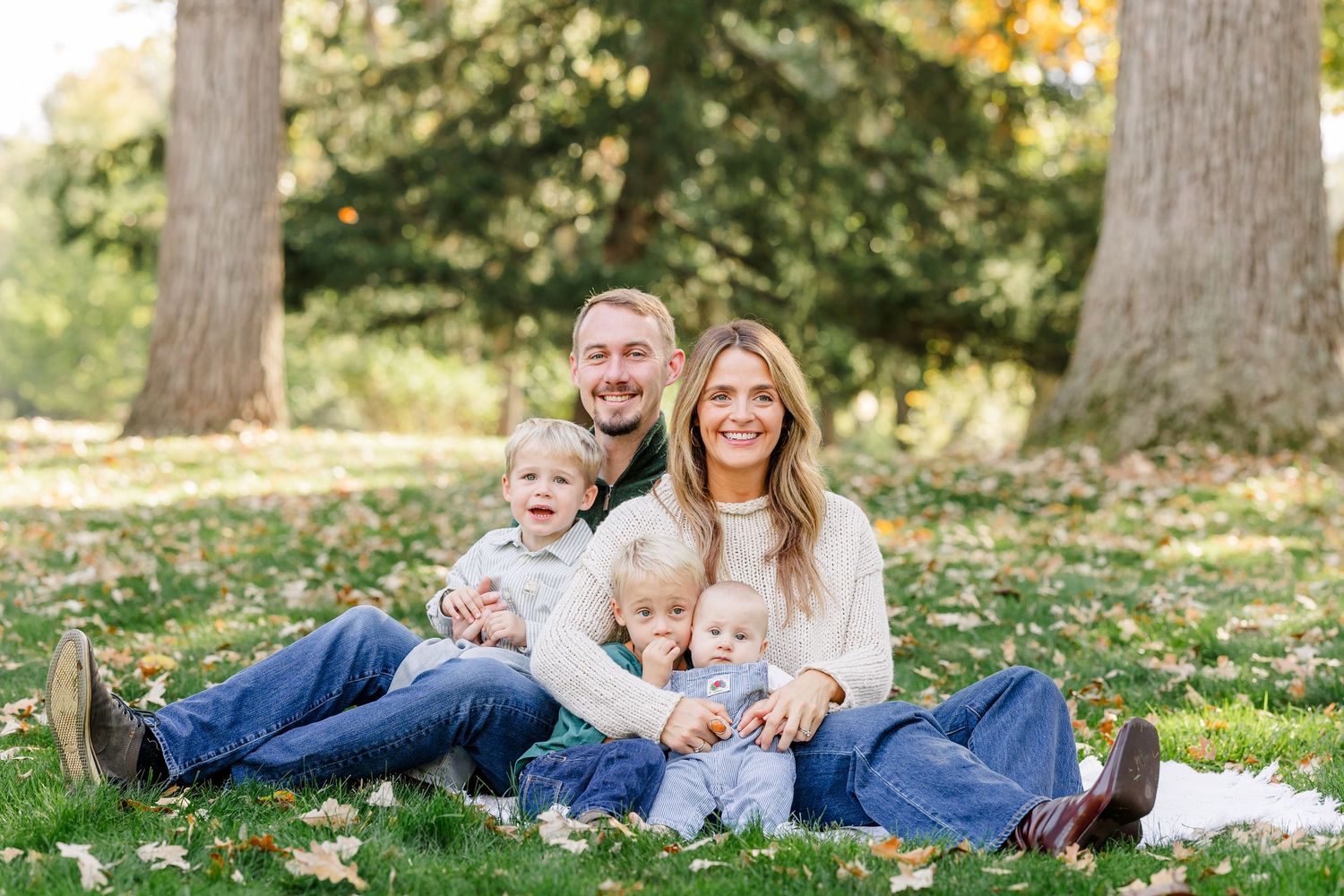 Family of five smiling outdoors on grass in a park, with two parents and three young children sitting together.