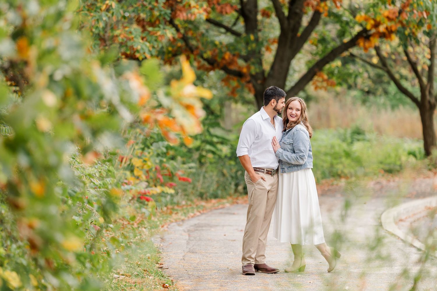 Couple in an embrace on a park path surrounded by colorful autumn foliage during an engagement photo session.
