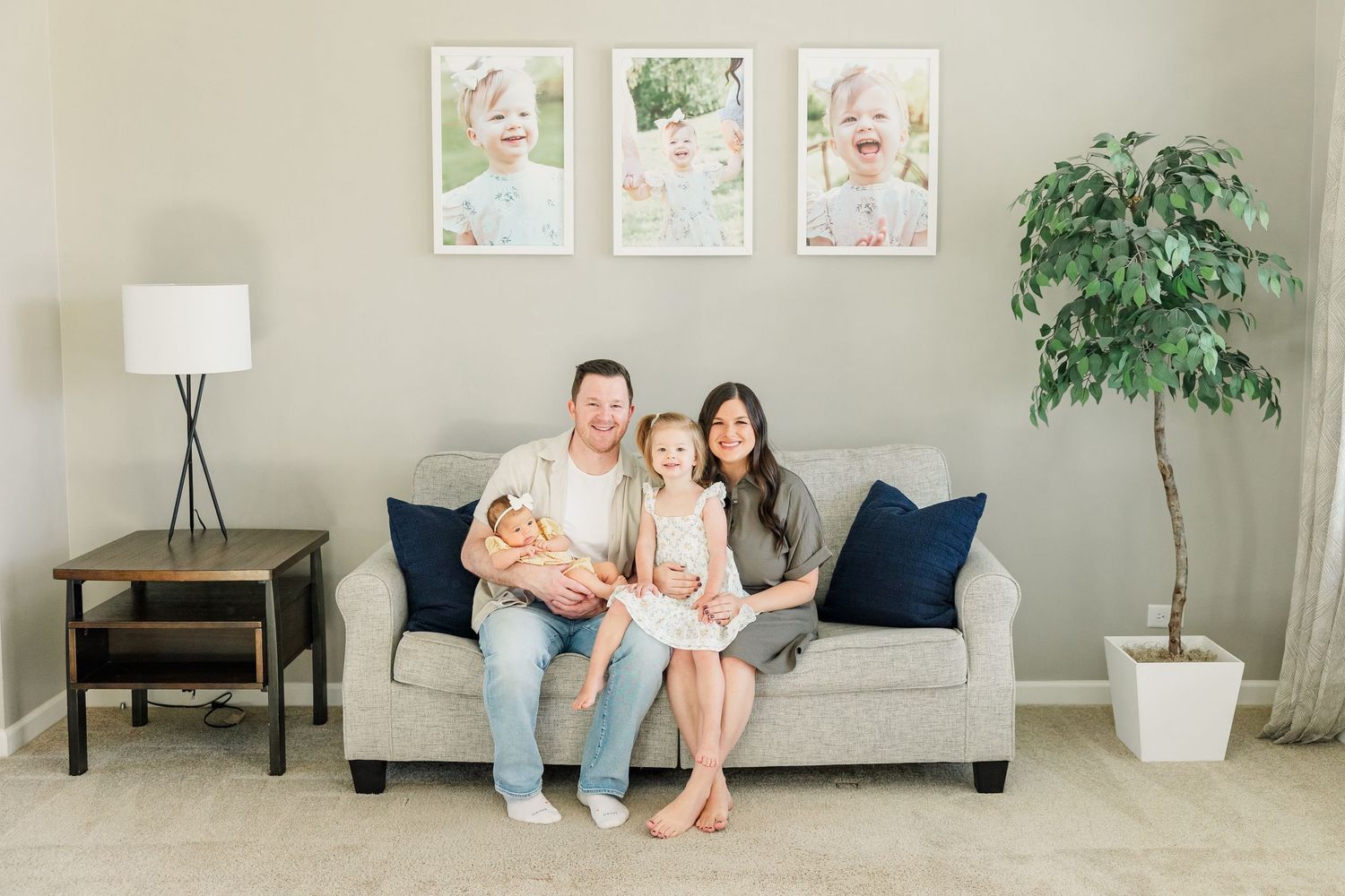 A happy family of four sits together on a gray sofa with blue pillows, framed children's photos hanging on the wall behind them.