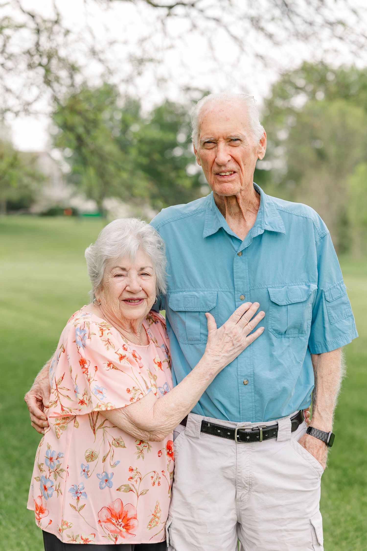 Smiling elderly couple posing outdoors on green grass, woman in floral pink top and man in blue shirt.