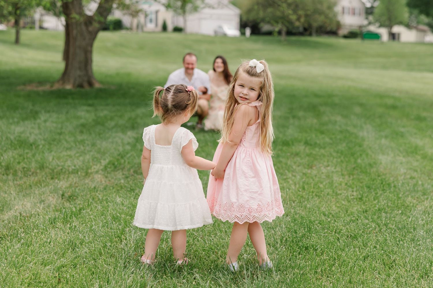 Two young girls in white and pink dresses hold hands on green lawn while parents watch with a dog in background.