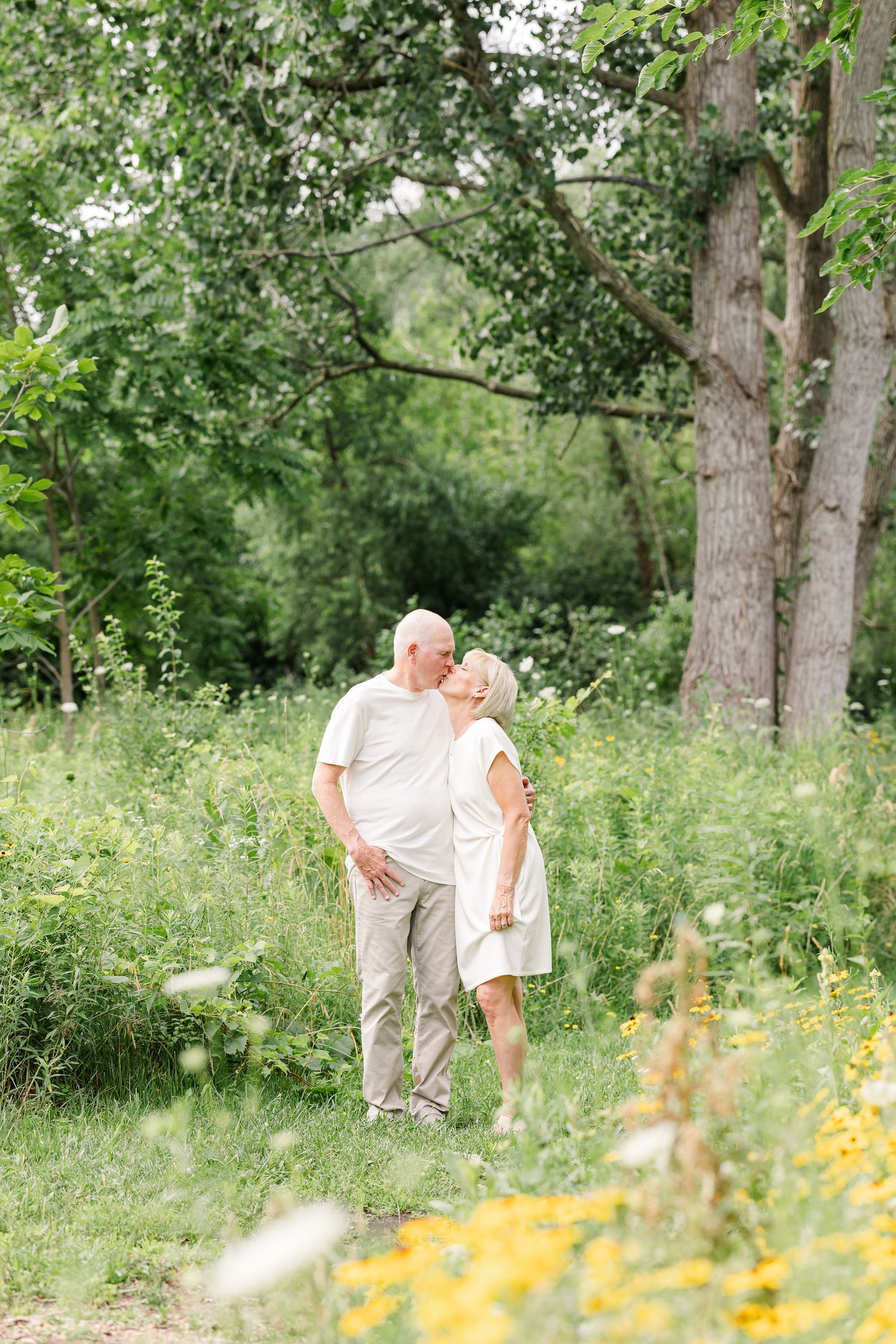 Elderly couple sharing a tender kiss in a lush green meadow with wildflowers and trees in summer.
