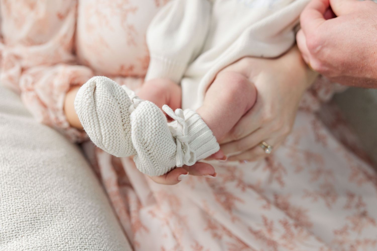 Adult hands gently holding tiny white knit baby booties on a newborn dressed in a floral pink outfit.