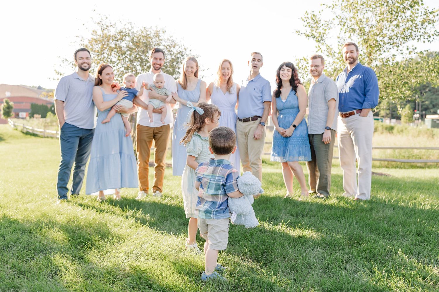 Large family group photo outdoors on green grass in soft golden light, with two small children running toward smiling adults.