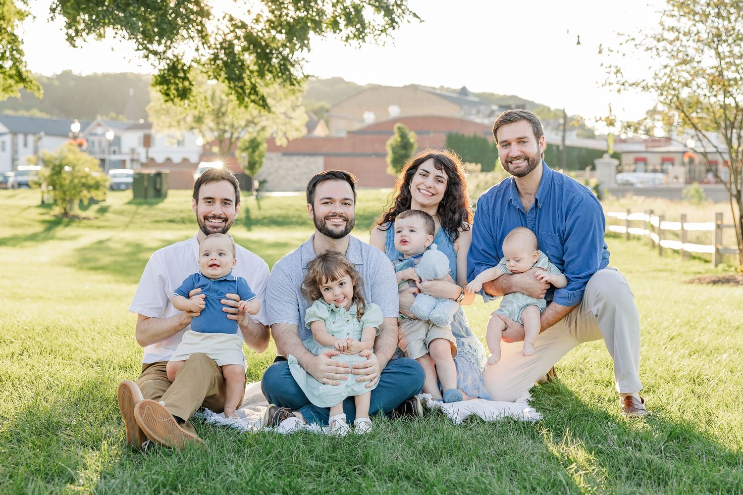 Two families with young babies and toddlers sit together smiling on green grass in a sunny park setting.
