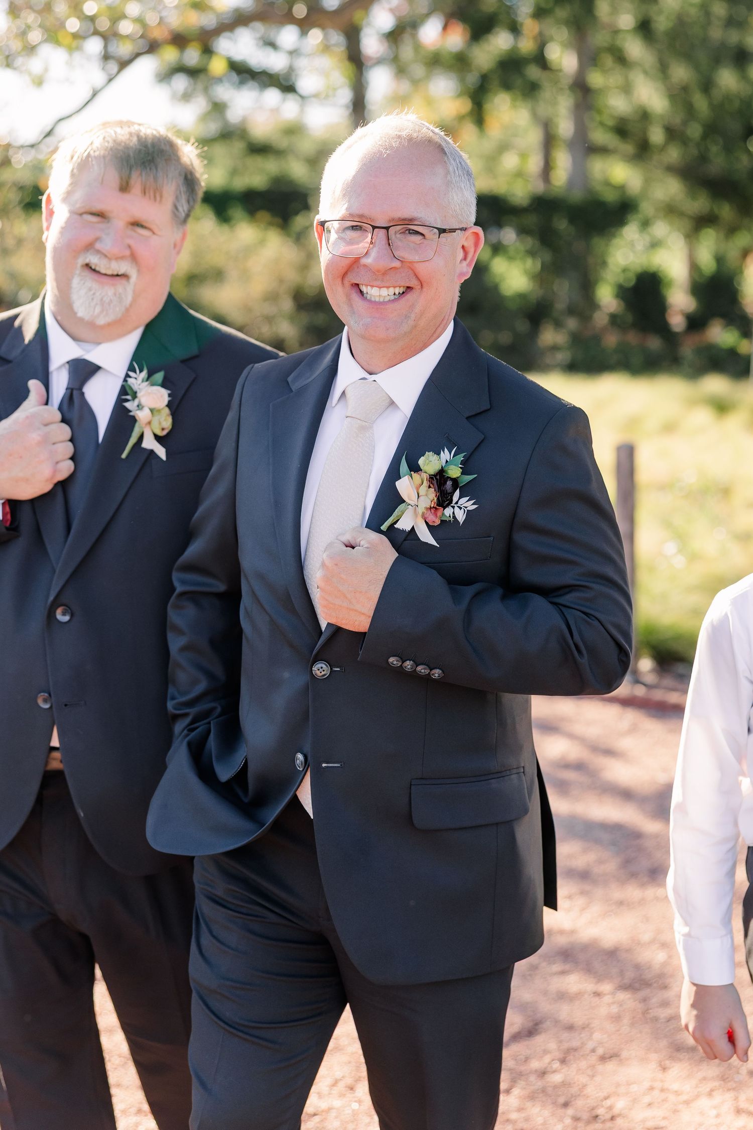 Two smiling men in dark suits with floral boutonnieres stand outdoors at a sunny wedding, one in glasses with a hand in his pocket.