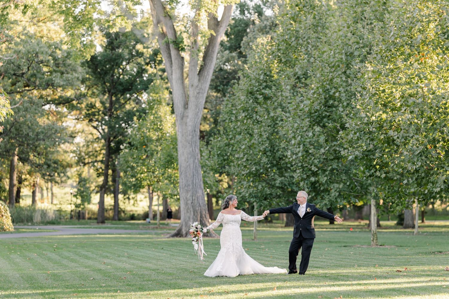Bride in a white gown and groom in a black suit hold hands and walk across a green lawn beneath tall trees at an outdoor wedding.