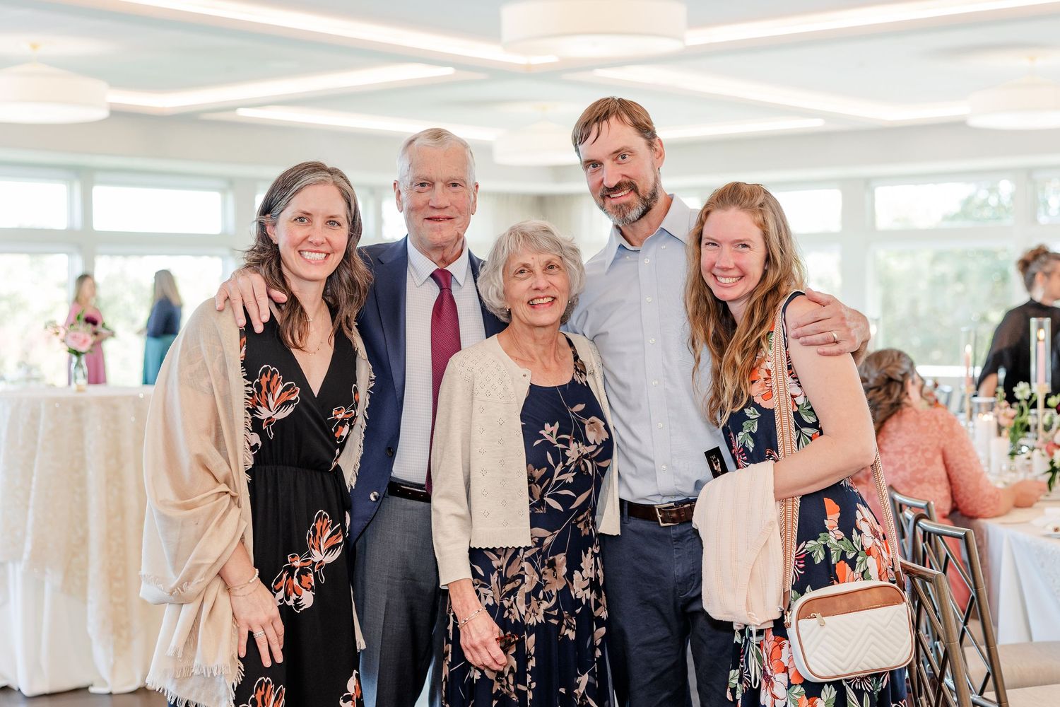 Five smiling family members in dressy attire pose with arms around each other in a bright wedding reception venue with guests behind.