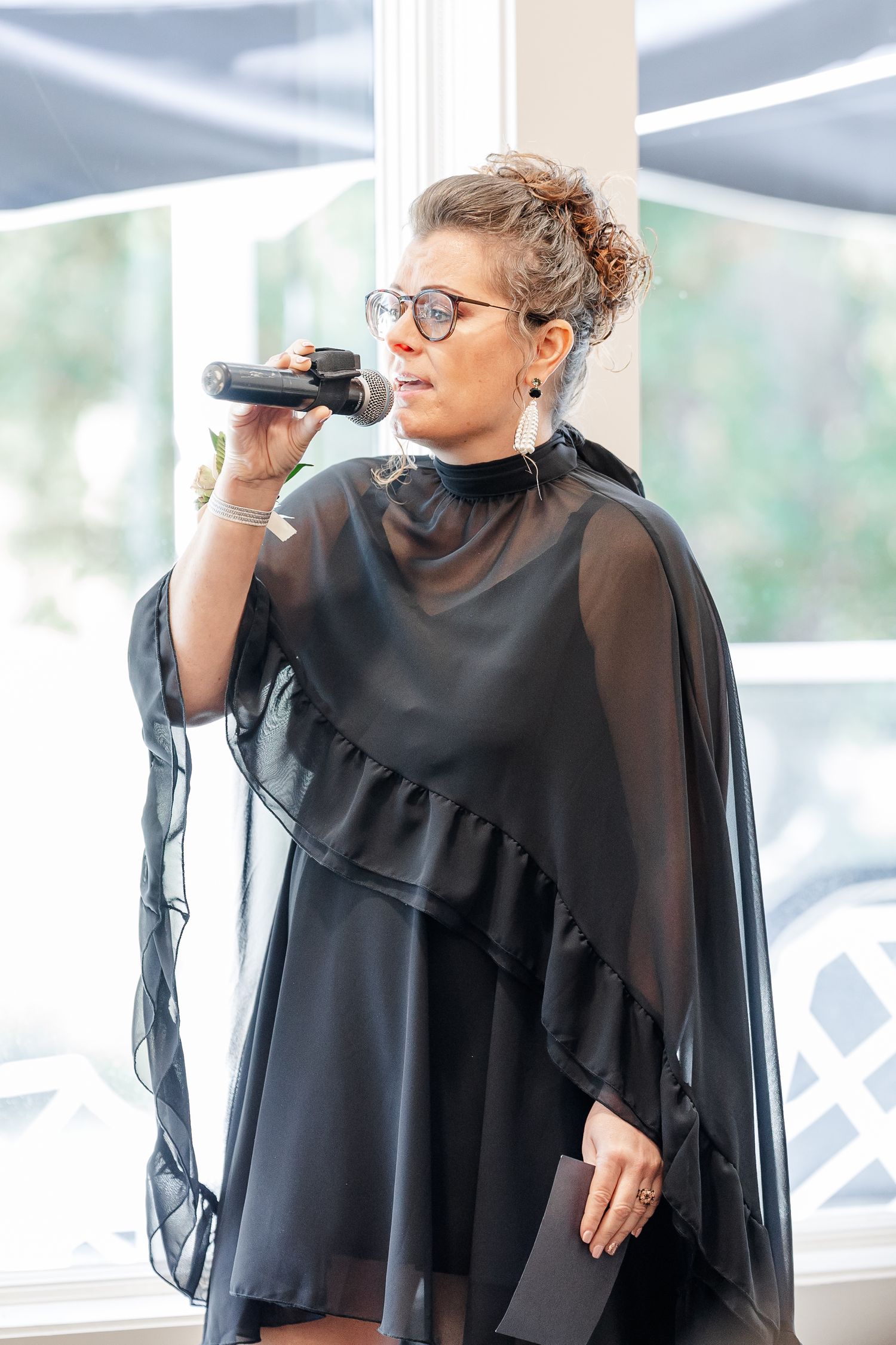 Woman with glasses and an updo speaks into a microphone at a wedding, wearing a black sheer cape dress and drop earrings.