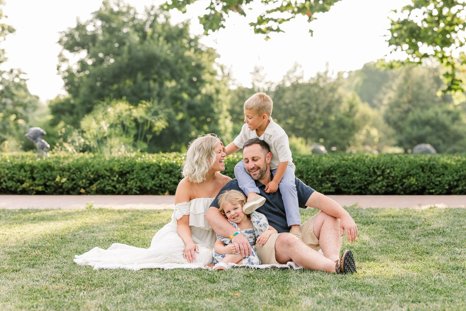 Family of four sitting on green grass in a sunny park, laughing and playing together outdoors.