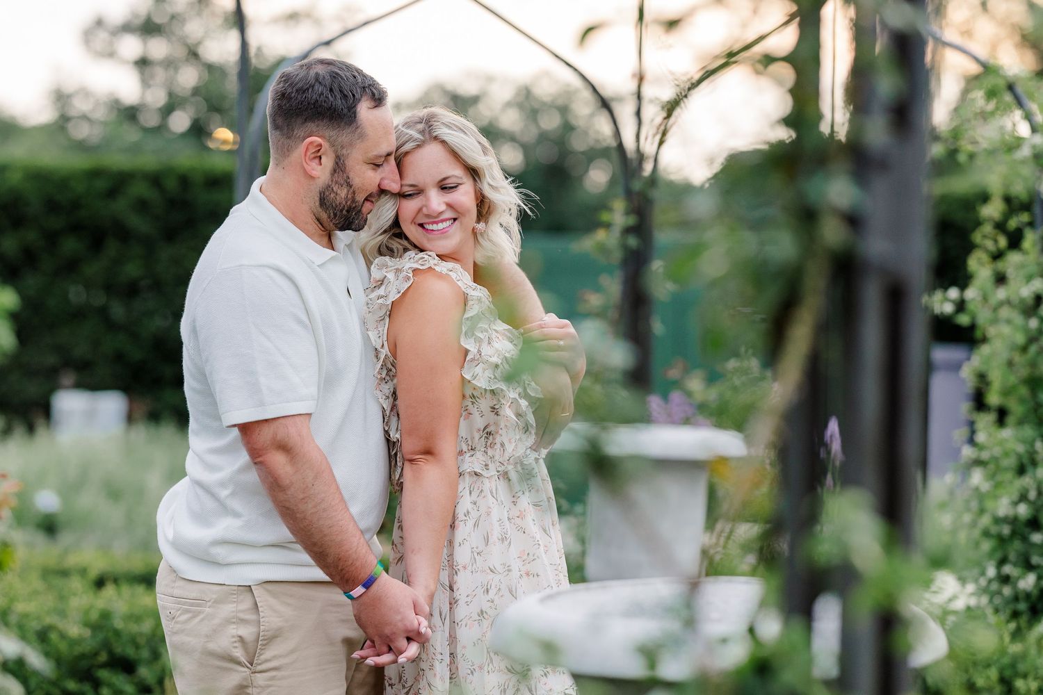 Couple shares romantic embrace in lush garden at sunset, woman smiling over shoulder at camera.