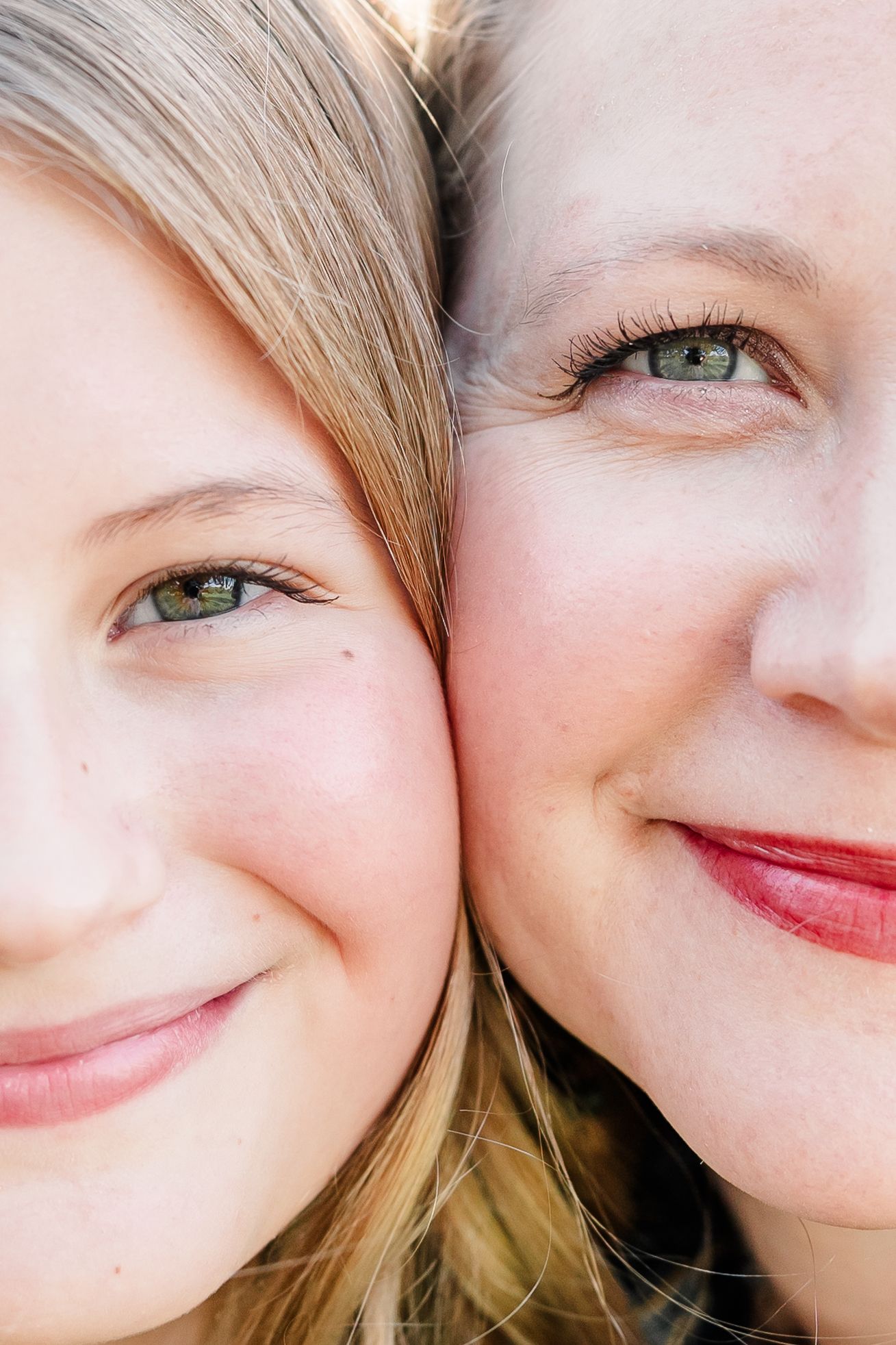 Close-up of two smiling faces pressed cheek to cheek, a young girl with blonde hair and a woman with green eyes and red lipstick.