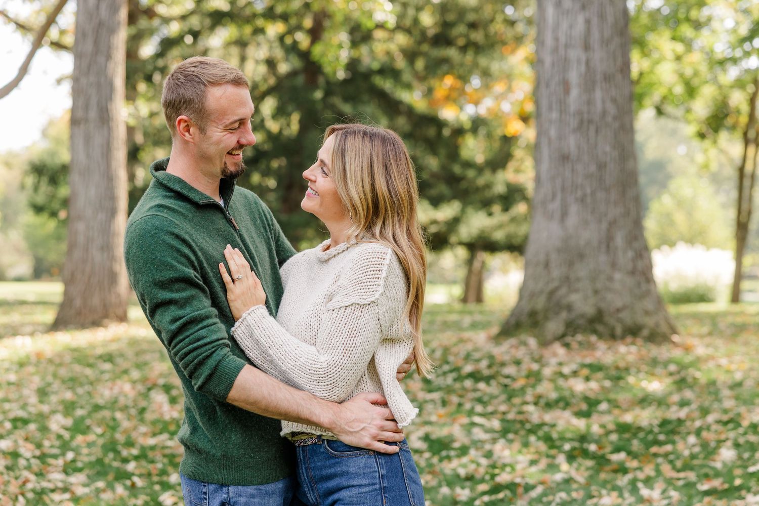 Couple smiling at each other while embracing in a sunlit park surrounded by fall foliage.