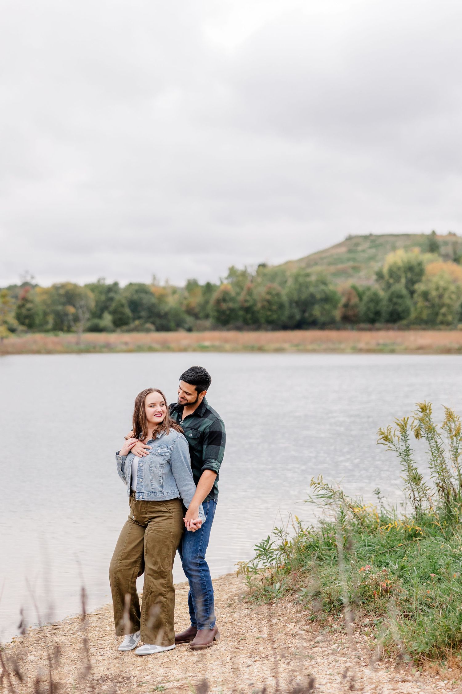 Smiling couple embracing on a lakeshore under an overcast sky, with a grassy hill and trees in the background.
