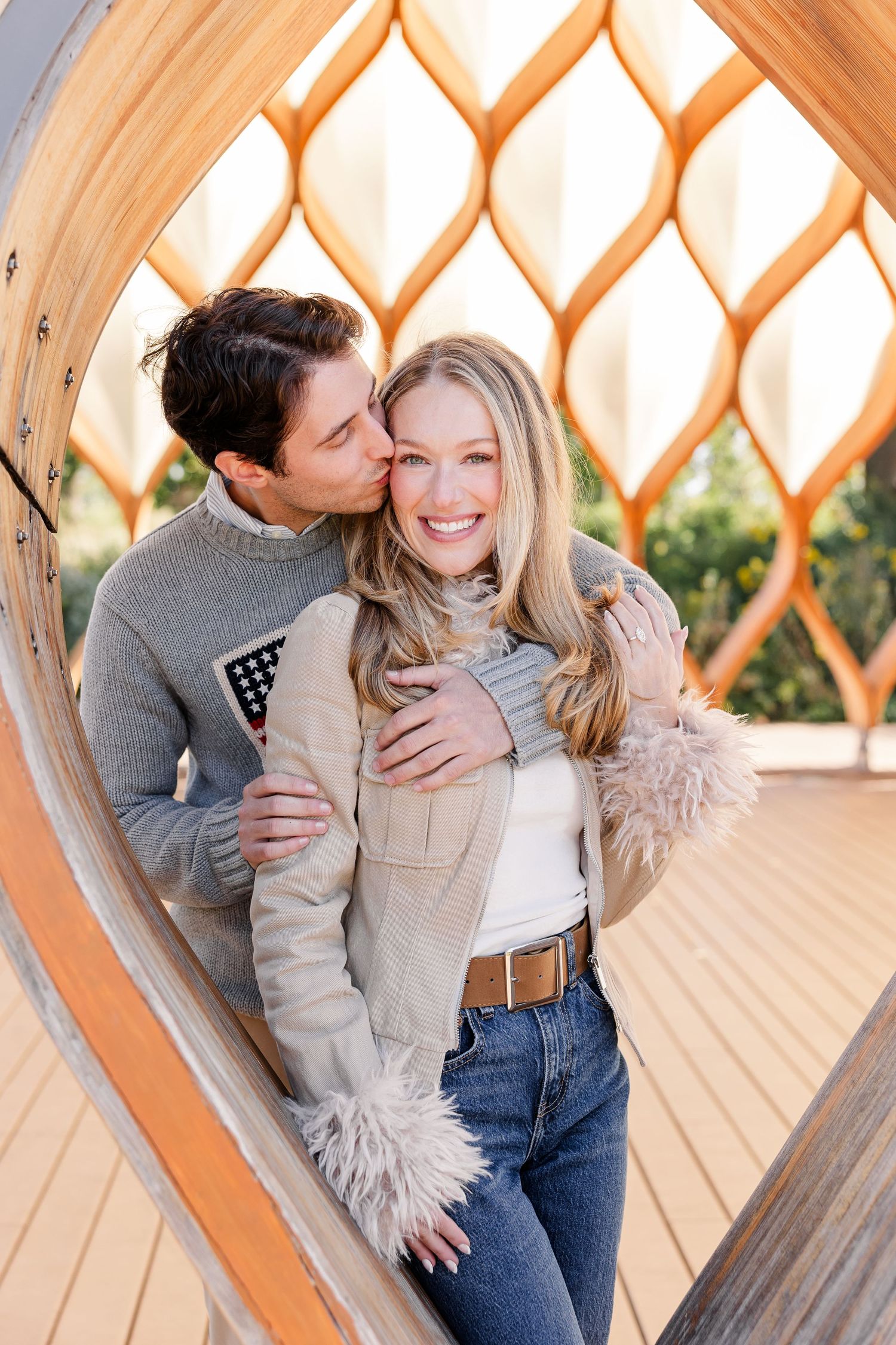 Couple embraces inside wooden lattice structure, man kisses womans cheek.