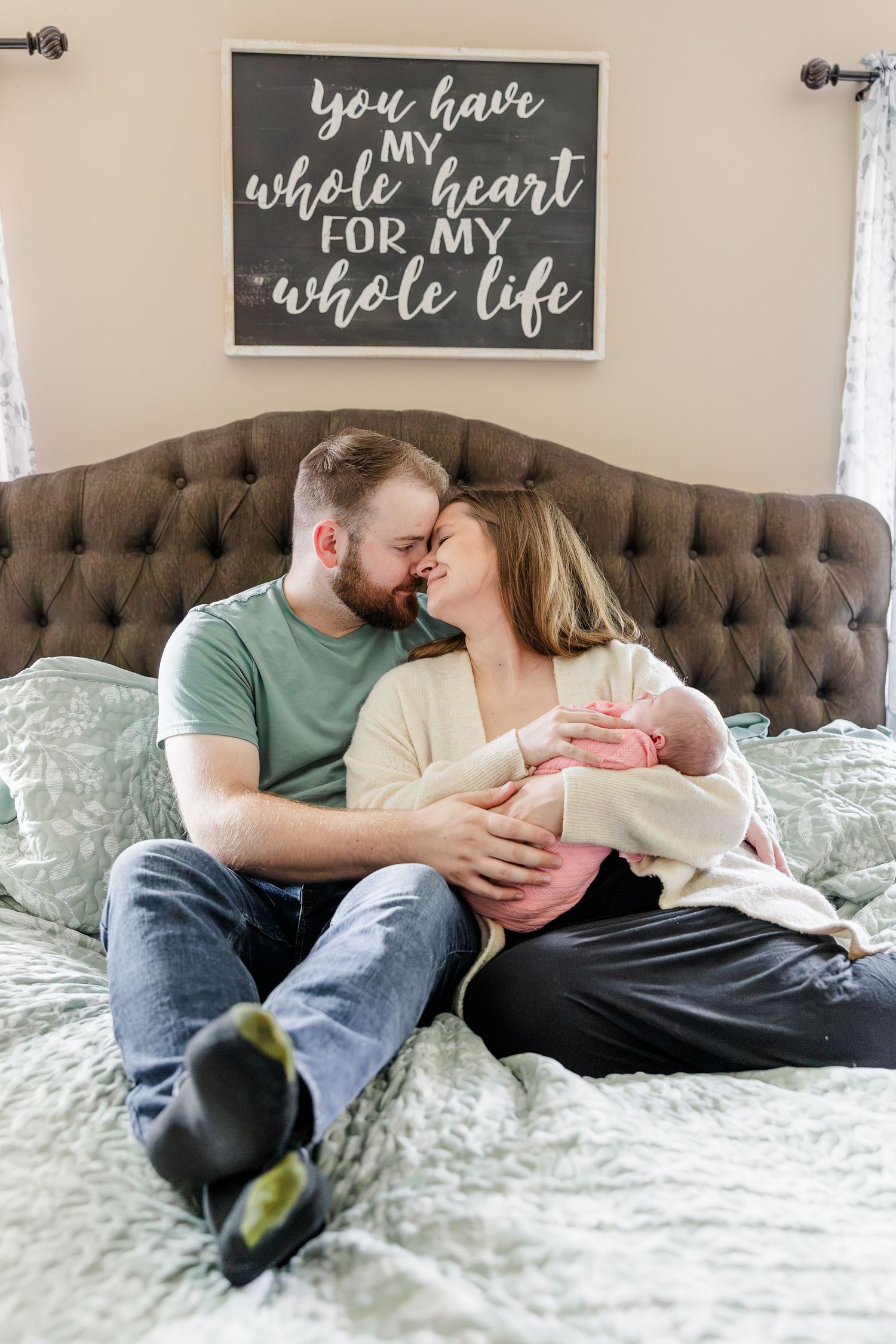 A couple shares a tender nose-to-nose moment on a bed while holding their newborn baby together.