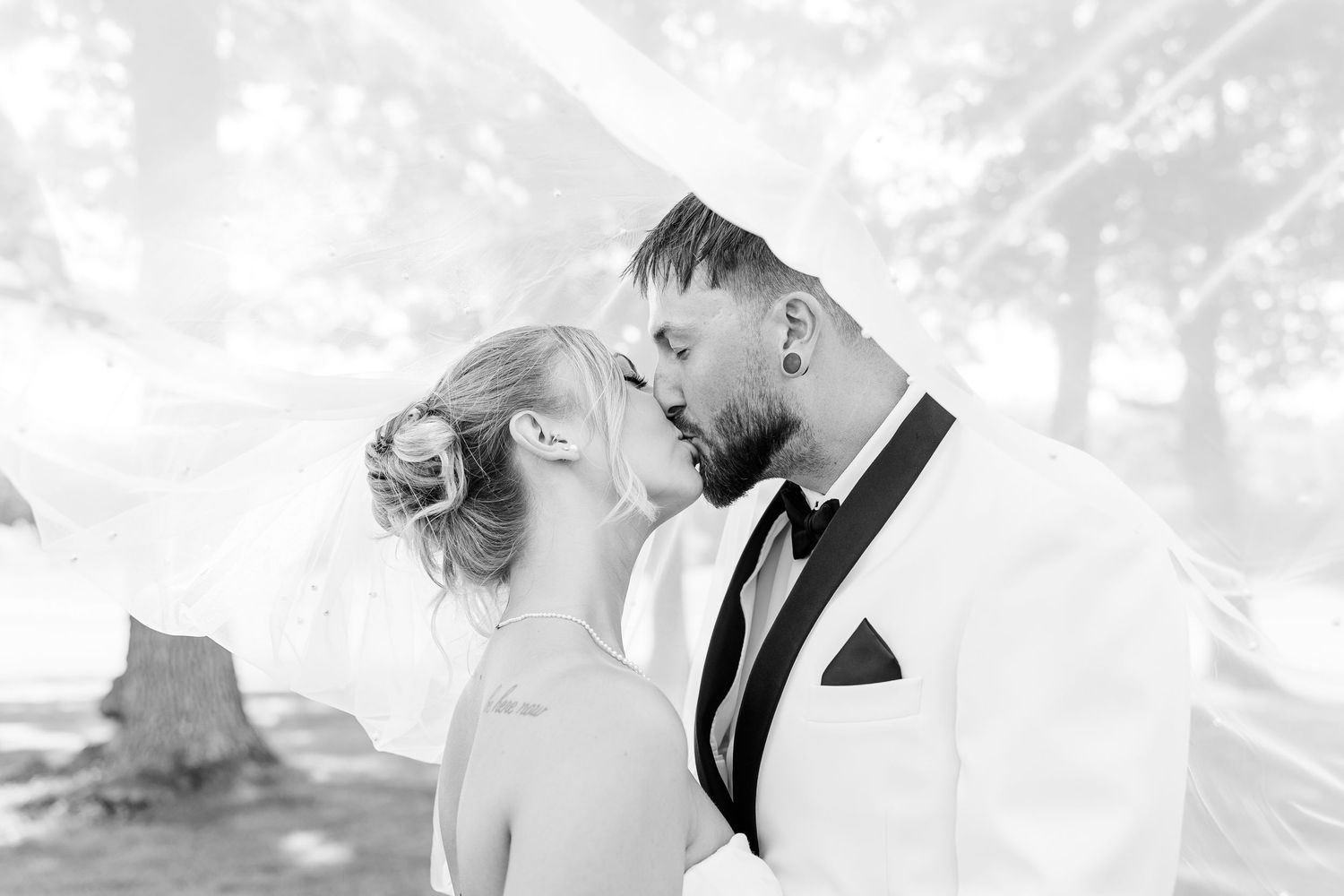 Black and white romantic kiss photo of bride and groom beneath a flowing veil outdoors.