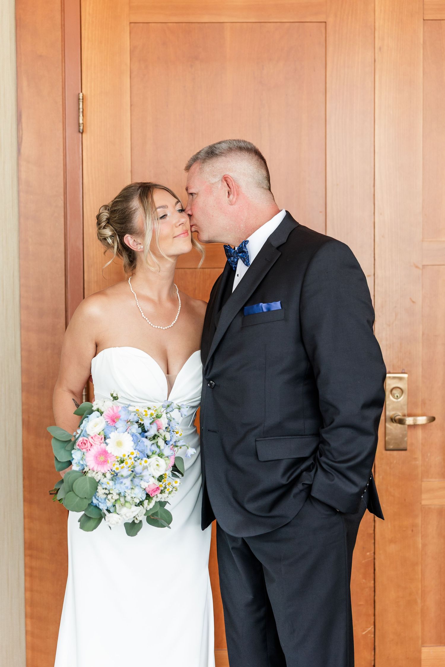 Groom in dark suit with blue bow tie kisses bride in white gown holding pastel floral bouquet by wooden door.