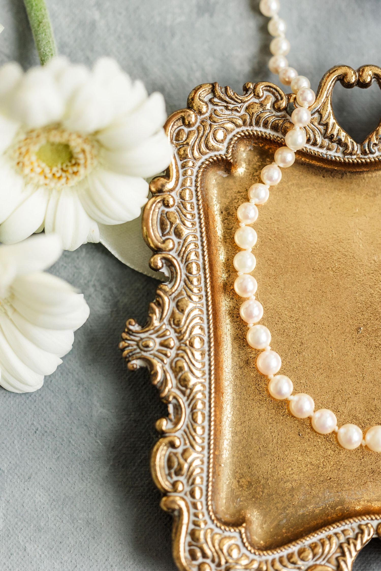 Vintage gold ornate tray with pearl necklace and white gerbera daisy on gray background.