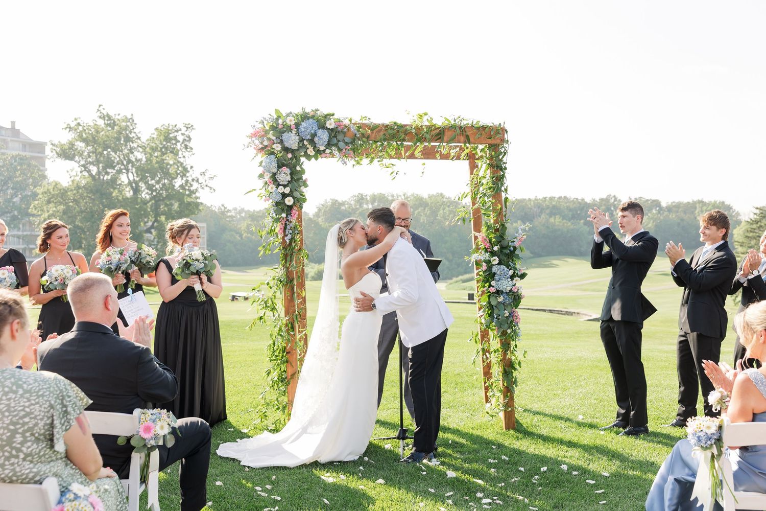 Bride and groom share first kiss under a floral arch on a green lawn during outdoor wedding ceremony.