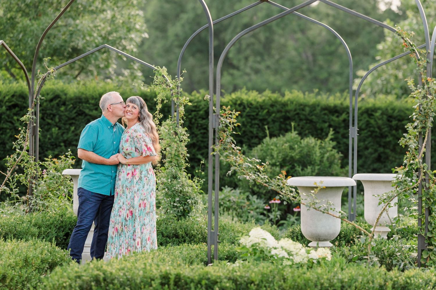 Couple embracing under a garden arbor surrounded by lush green hedges and roses during an outdoor engagement session.
