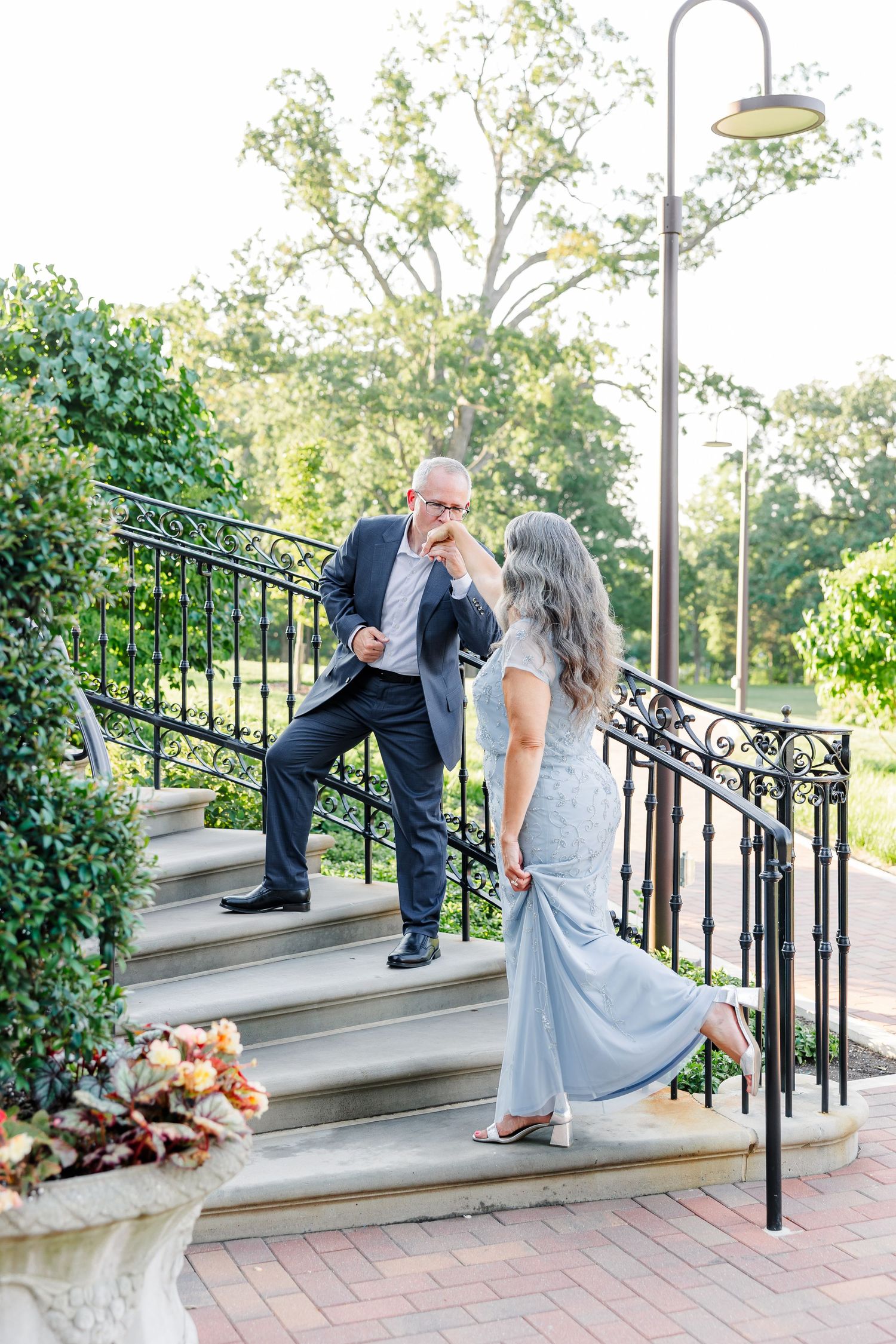 A man in a navy suit kisses a woman's hand on elegant outdoor stone steps at a wedding venue.