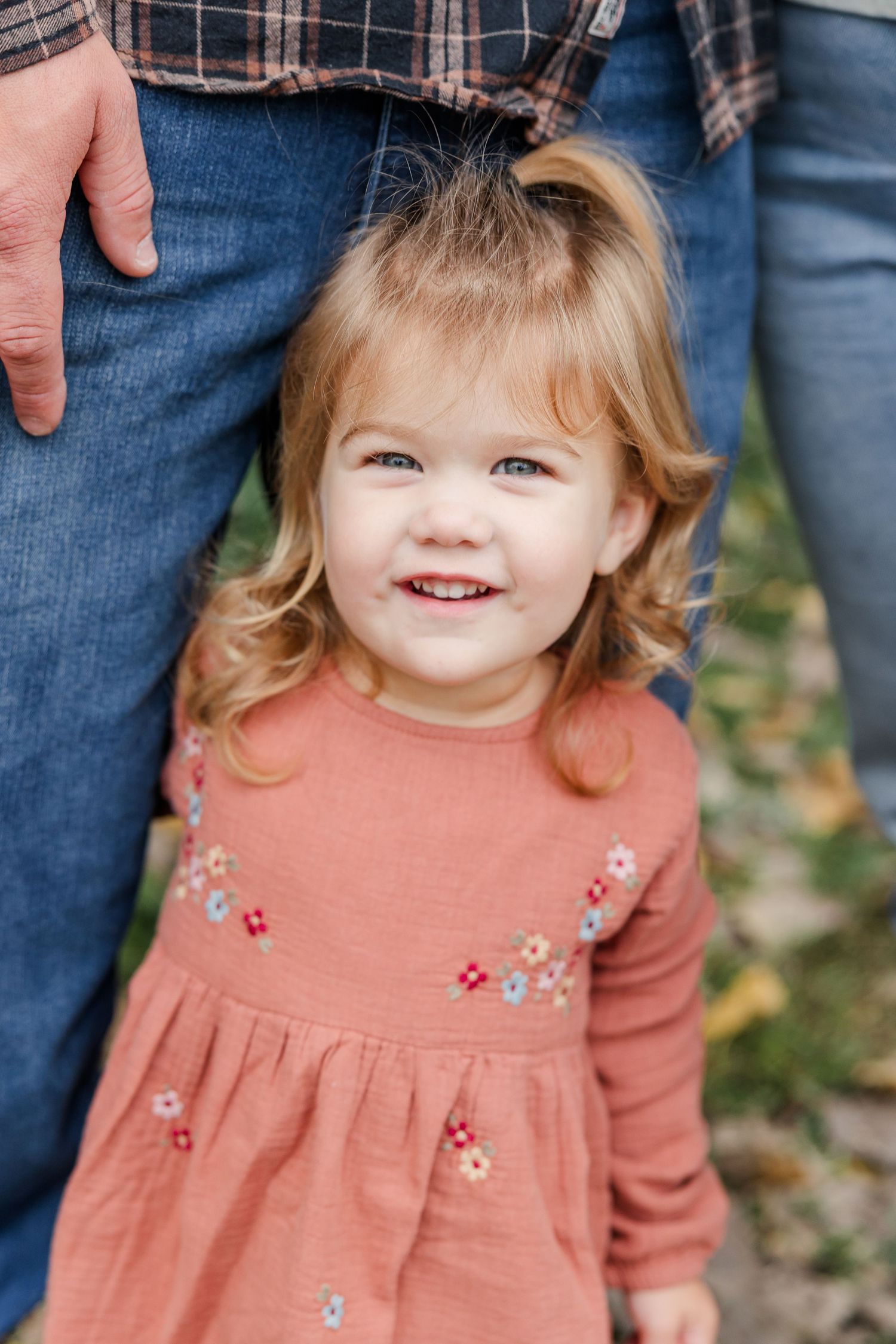 Adorable toddler girl in a pink floral dress smiles brightly while standing beside her father outdoors.