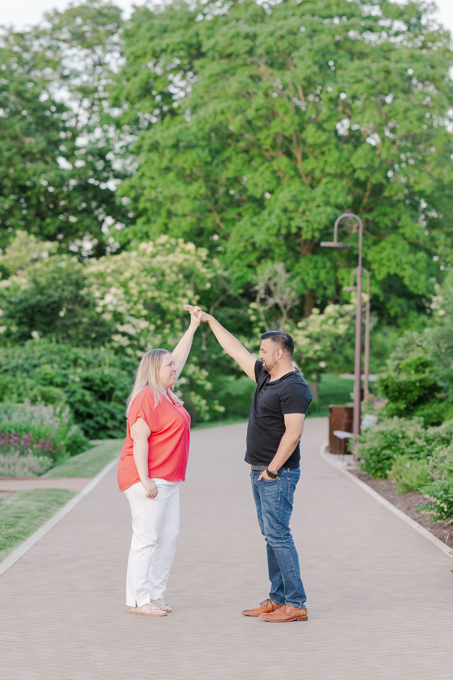 Couple sharing playful dance-like pose on garden pathway surrounded by lush trees.