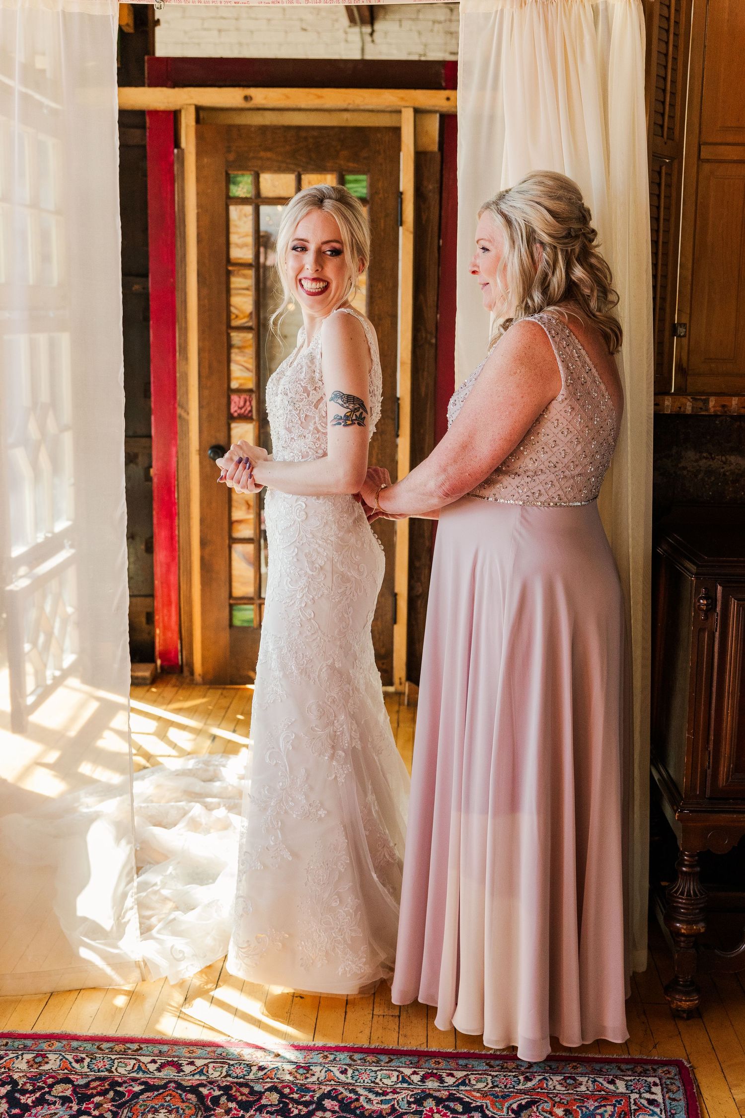 Two women sharing a joyful moment in a sunlit room, wearing formal dresses in pink and white lace.