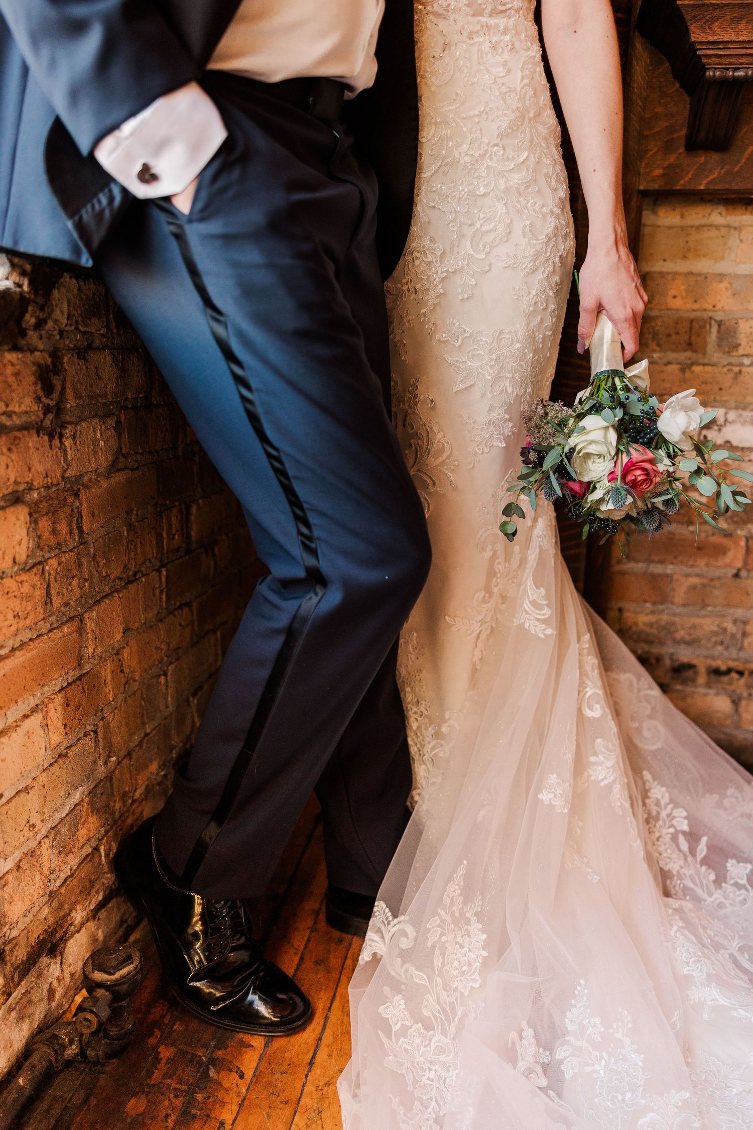 Detail shot of wedding couple's legs and dress hem against rustic brick wall with floral bouquet.