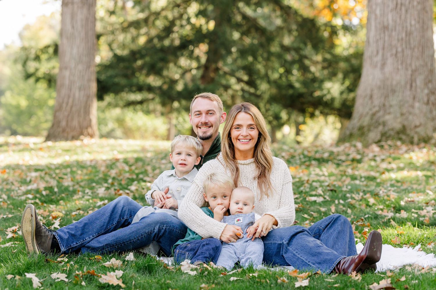 Family of four smiling outdoors in a park during autumn, sitting on grass with large trees and fallen leaves.