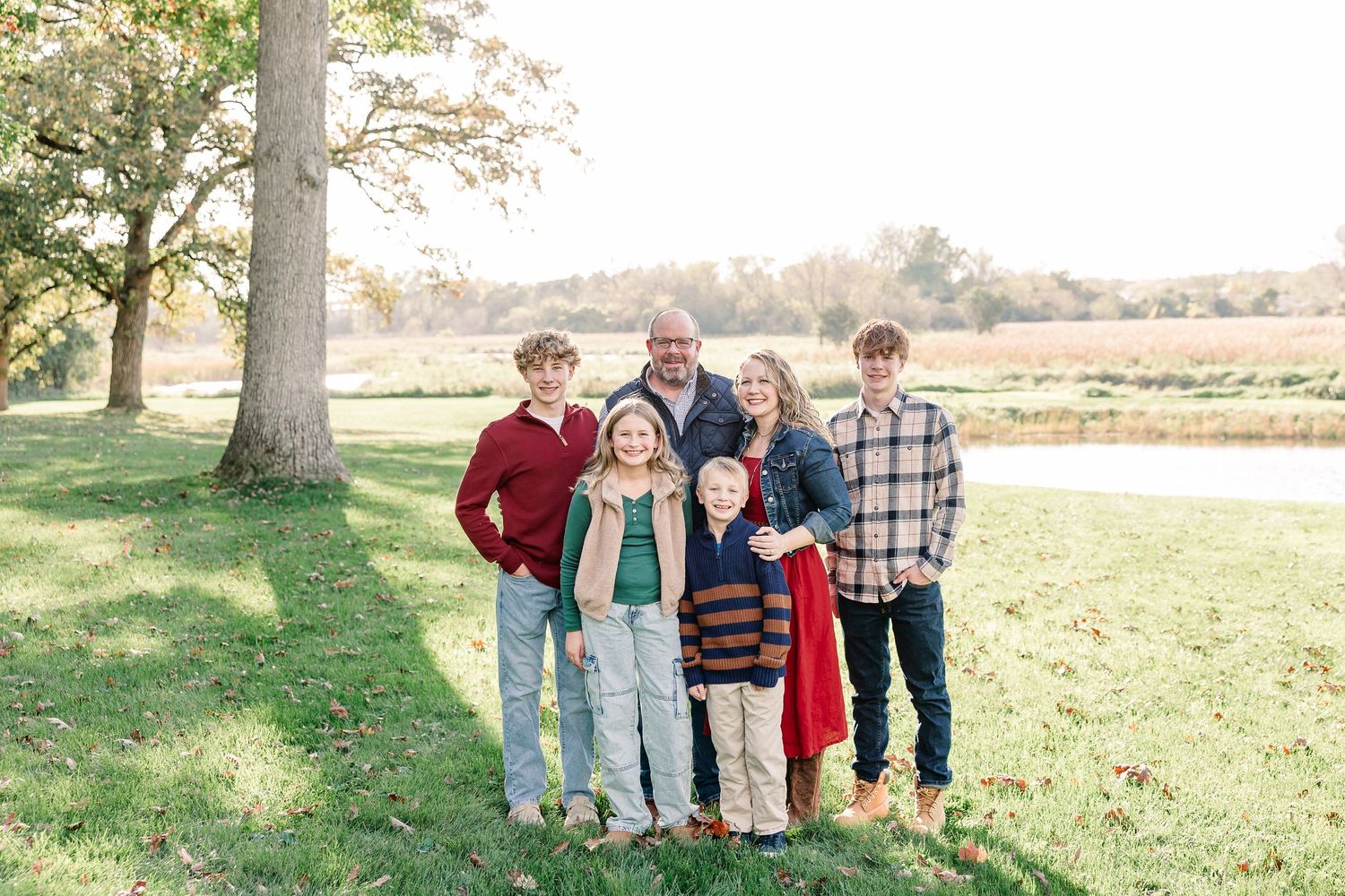 Family of six poses together on a sunny grassy lawn beside a large tree, with a pond and open field in the background.