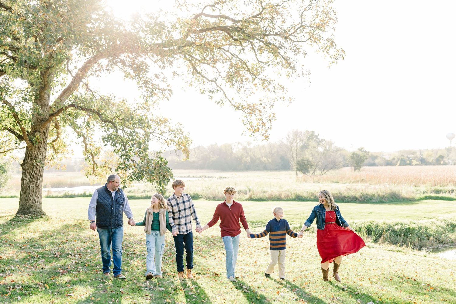 A family of six holds hands in a line while walking across a sunlit grassy field beneath a large tree during an autumn family photo session.