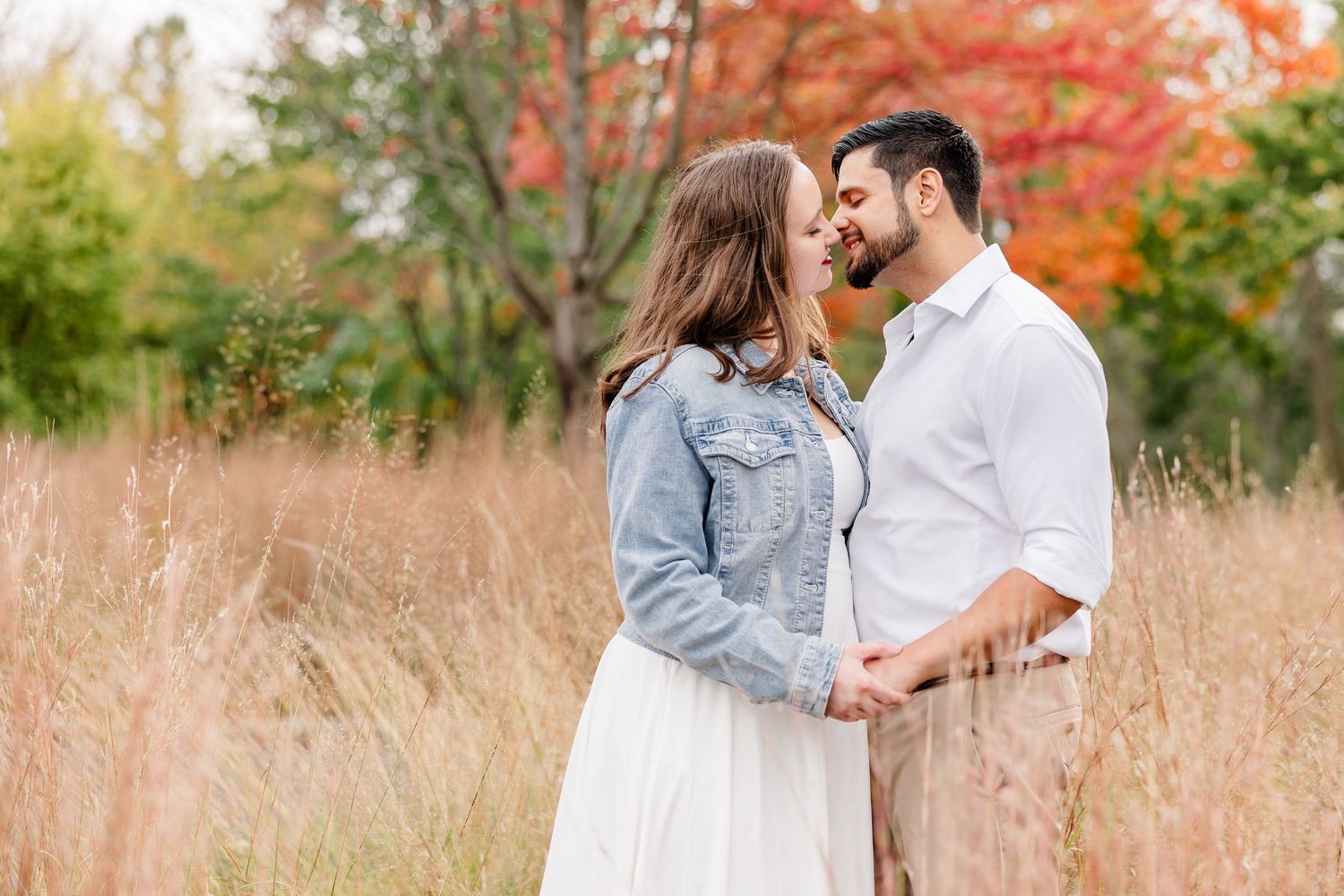 Couple in a denim jacket and white shirt share a close, romantic moment in a golden grass field with autumn trees behind.