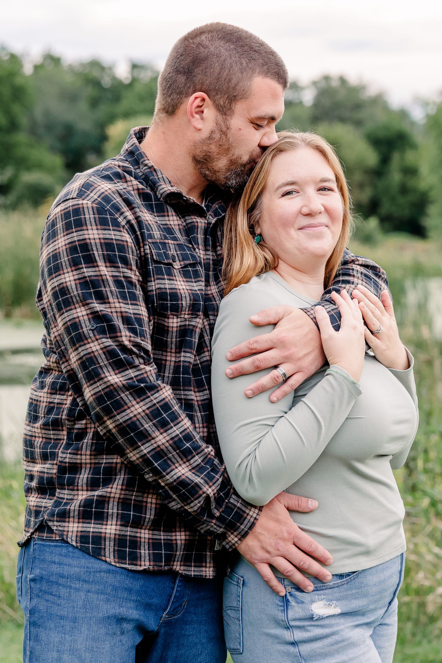 A man in a plaid shirt embraces and kisses a smiling woman near a pond in an outdoor setting.