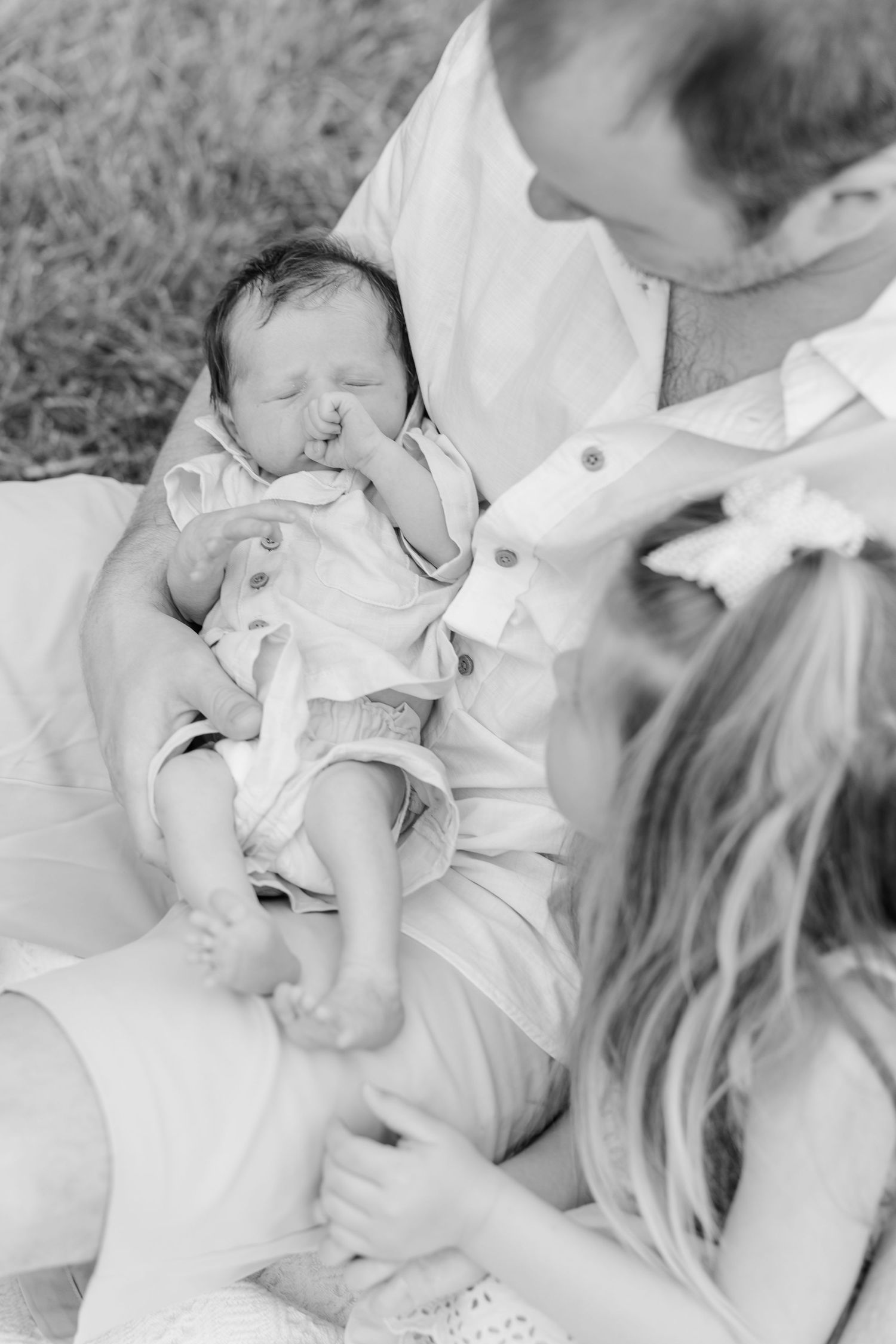 Black and white photo of parents and siblings tenderly gathered around a sleeping newborn baby on a blanket outdoors.
