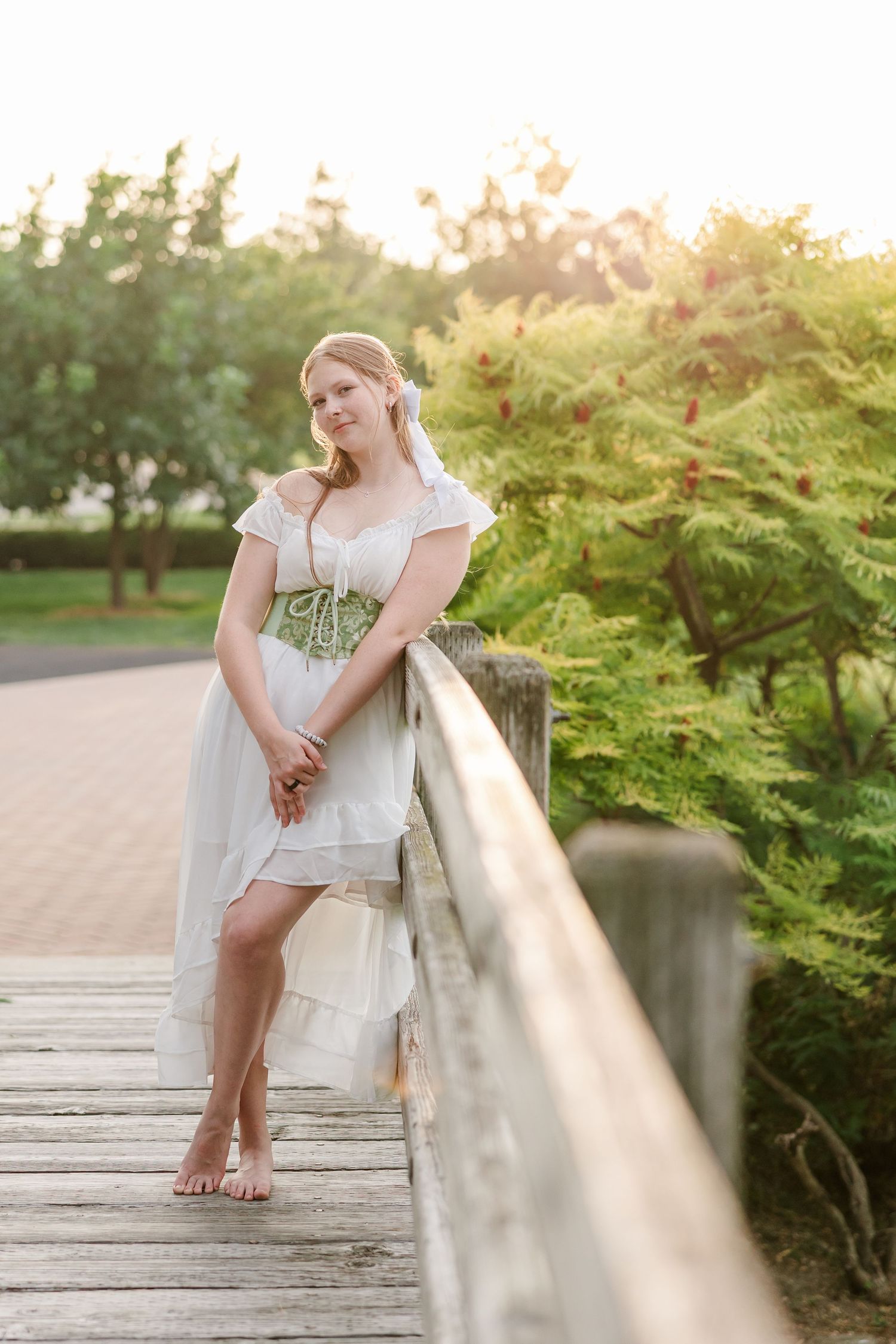 Golden-hour senior portrait of Samantha in a white dress and green corset beside a rustic garden bridge.